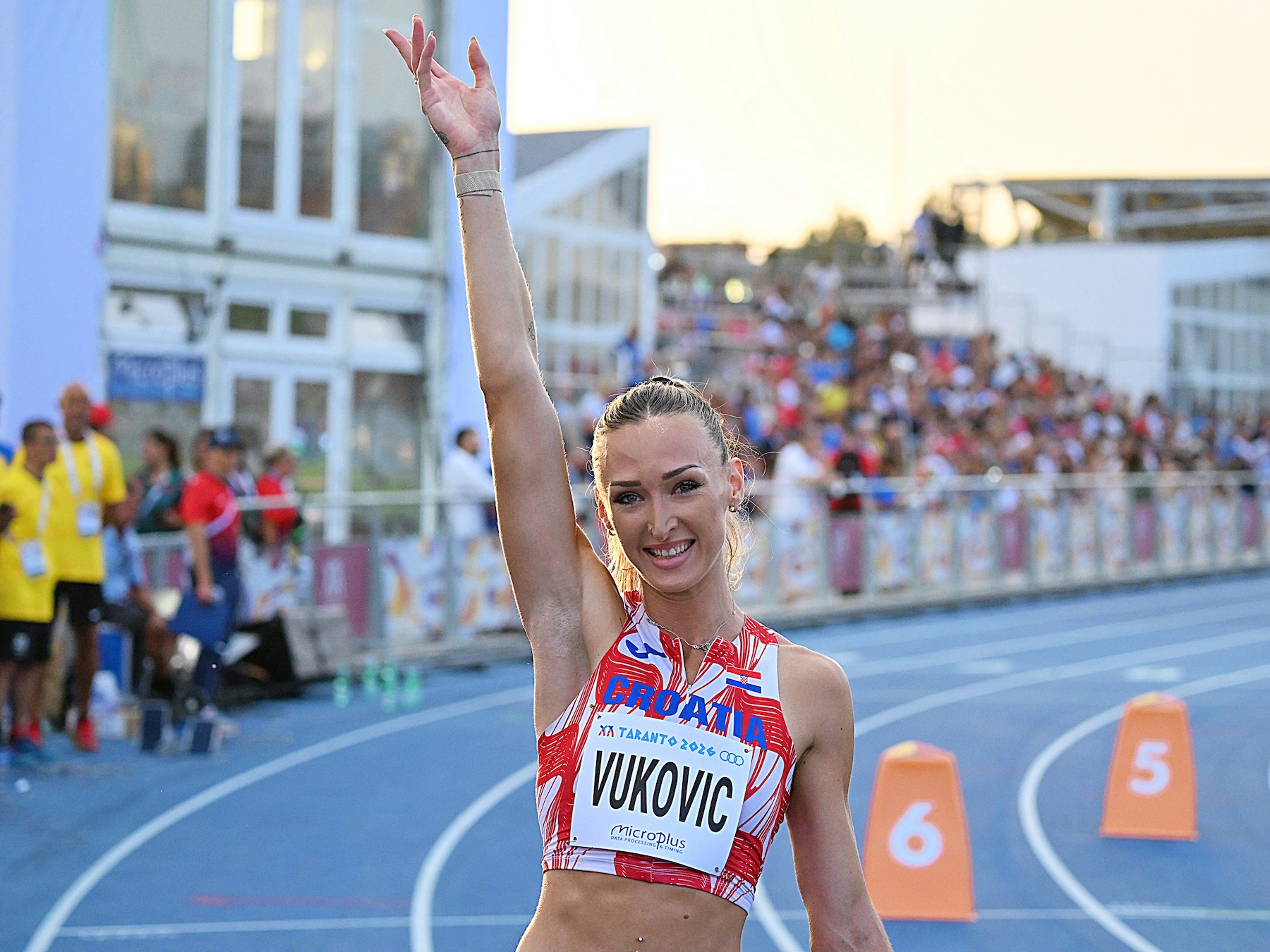 An athlete in a red and white Croatia uniform raises one arm in celebration on a blue track, with spectators visible in the background.